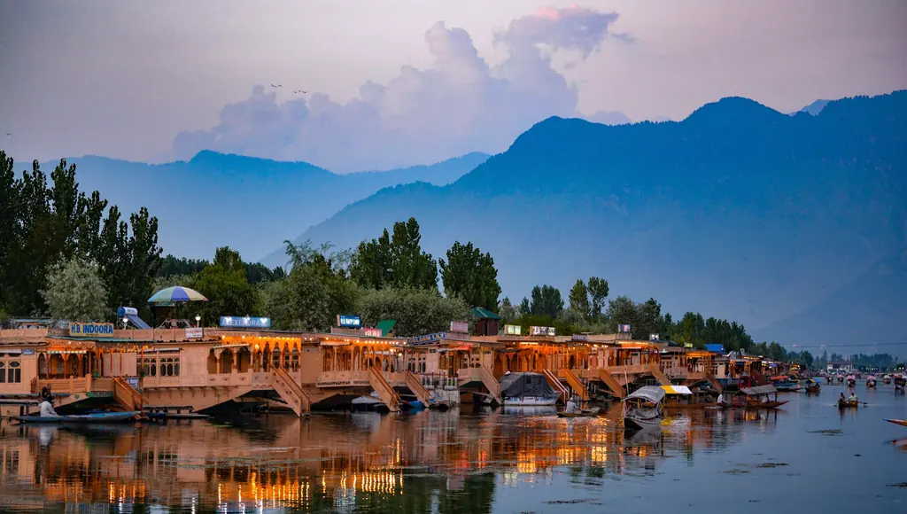 Rows of carved houseboats lining Dal Lake in Srinagar at dusk, their lights reflected beneath blue Zabarwan hills