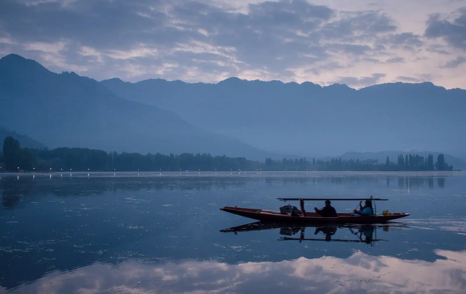 A shikara with its boatman and guests gliding across a still Dal Lake at first light, the Zabarwan hills in blue morning haze